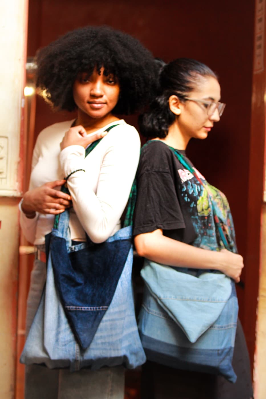 Two women standing back to back at a doorway, each carrying a SamZar denim shoulder bag.