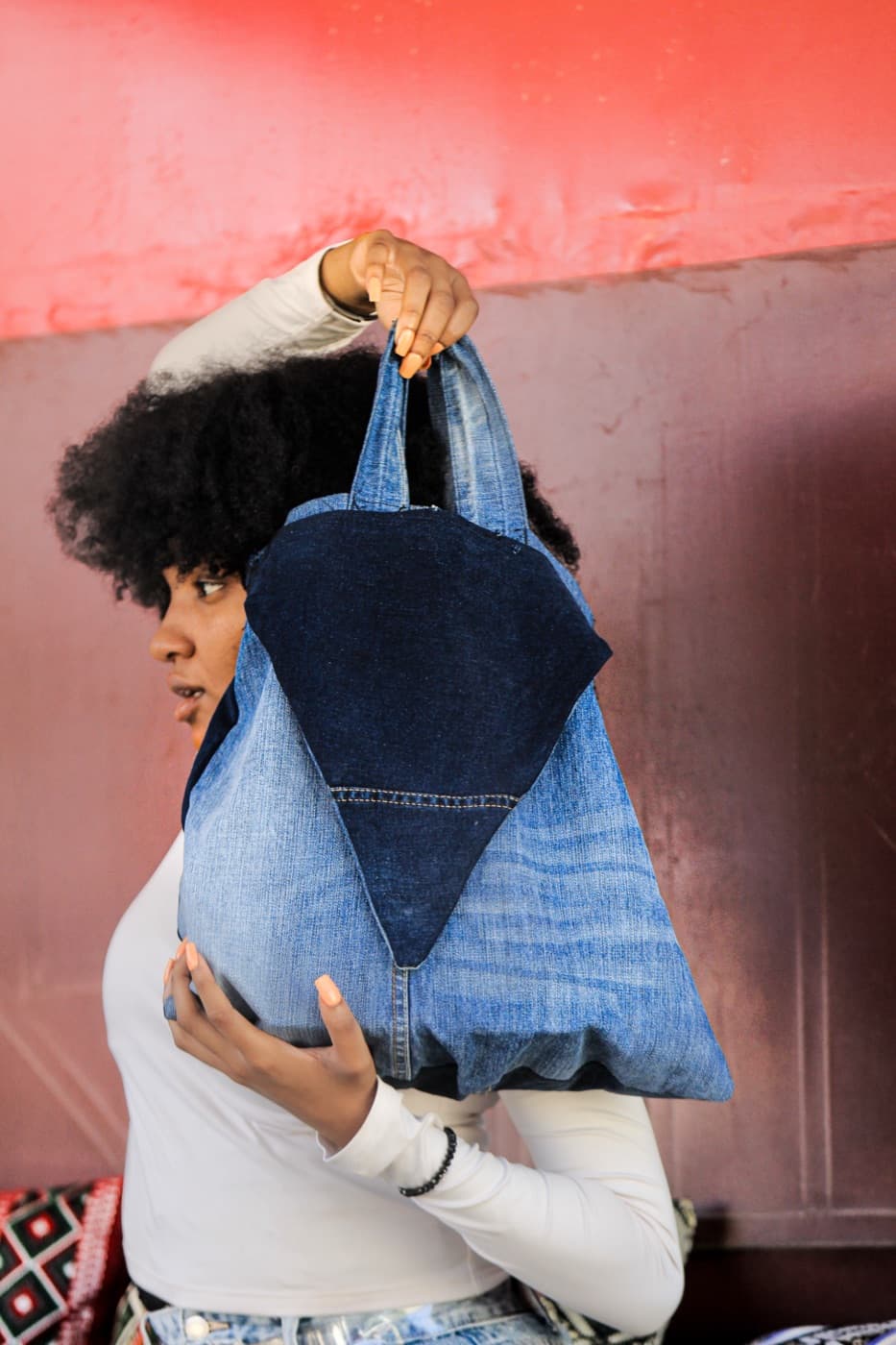 A woman in profile against a pink wall, lifting a SamZar denim bag over her shoulder.